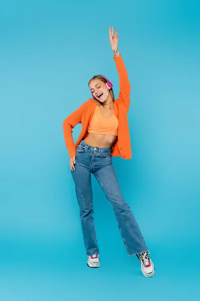 Pleine longueur de femme joyeuse dans les écouteurs chantant sur fond bleu — Photo de stock