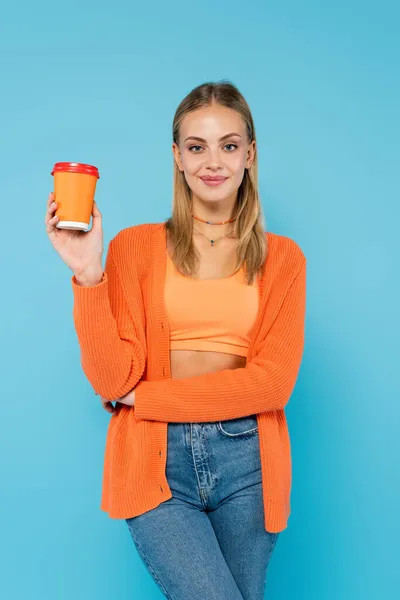 Mujer rubia bonita con taza de papel mirando a la cámara aislada en azul - foto de stock