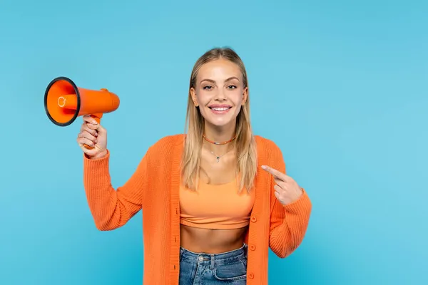 Smiling blonde woman pointing at orange loudspeaker isolated on blue — Stock Photo