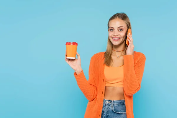 Mujer rubia sonriente sosteniendo una taza de papel y hablando en un teléfono inteligente aislado en azul - foto de stock