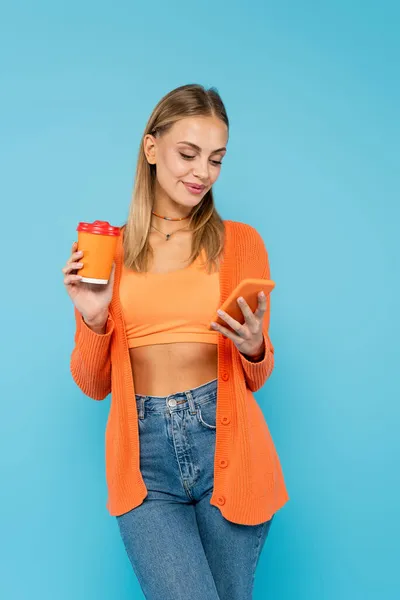 Mulher loira bonita usando smartphone e segurando copo de papel isolado no azul — Fotografia de Stock