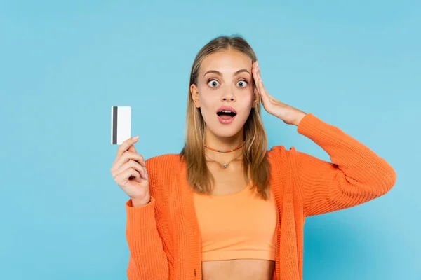 Shocked blonde woman holding credit card isolated on blue — Stock Photo