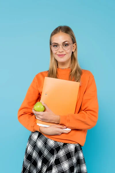 Студент блондинки в окулярах тримає паперову теку і яблуко ізольовано на синьому — стокове фото