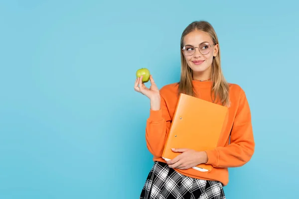 Estudiante positivo en anteojos con carpeta de papel y manzana aislada en azul - foto de stock