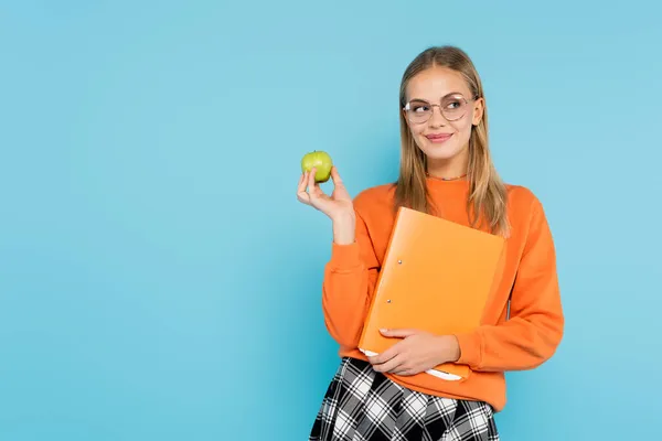 Studente positivo in occhiali in possesso di cartella di carta e mela isolato su blu — Foto stock