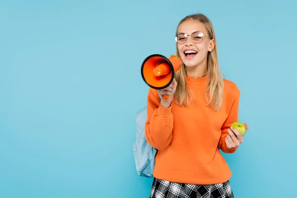 Blonde Studentin mit Brille schreit in Lautsprecher und hält Apfel isoliert auf blauem Grund — Stockfoto