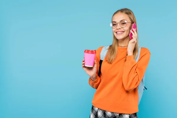 Estudiante sonriente en anteojos hablando por celular y sosteniendo café para ir aislado en azul - foto de stock