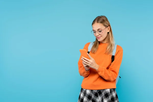 Blonde Studentin mit Rucksack und Brille, Handy isoliert auf blauem Grund — Stockfoto