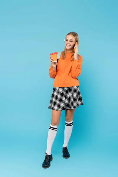 Full length of student in skirt holding paper cup and talking on mobile phone on blue background — Stock Photo