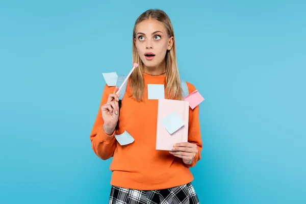 Pensive student in sticky notes holding pen and notebook isolated on blue — Stock Photo