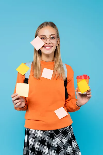 Estudiante sonriente en notas adhesivas sosteniendo una taza de papel aislada en azul - foto de stock