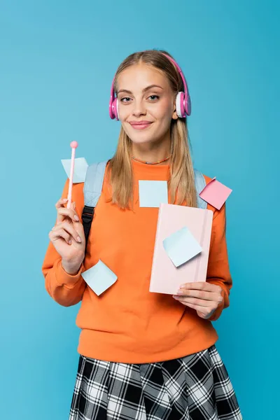 Graziosa studentessa in cuffia e appiccicosa appunti con taccuino e penna isolati su blu — Foto stock