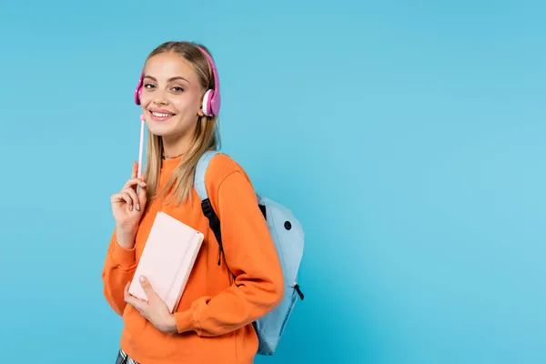 Веселий студент в навушниках тримає блокнот і ручку ізольовано на синьому — стокове фото