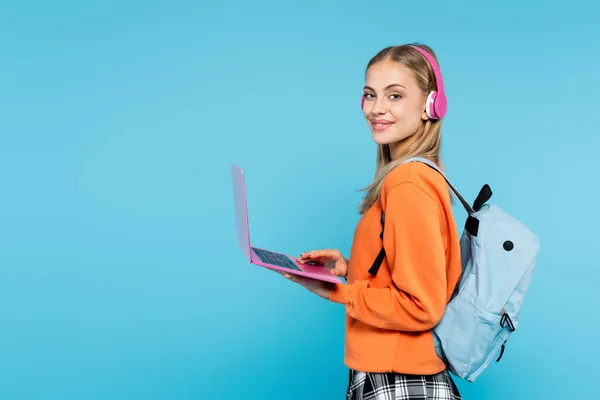 Blonde student in headphones and backpack holding laptop isolated on blue — Stock Photo