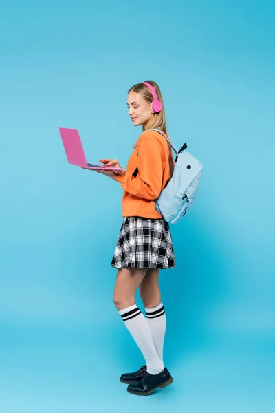 Comprimento total de estudante loira em fones de ouvido usando laptop no fundo azul — Fotografia de Stock