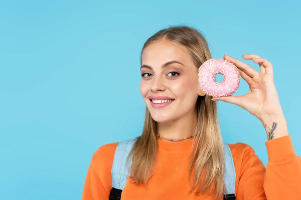 Estudiante feliz en sudadera naranja sosteniendo donut y mirando a la cámara aislada en azul - foto de stock