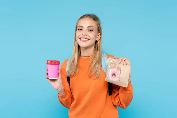 Cheerful student holding donut in package and takeaway drink isolated on blue — Stock Photo