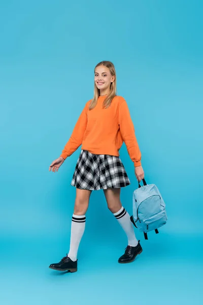 Full length of smiling blonde student carrying backpack on blue background — Stock Photo