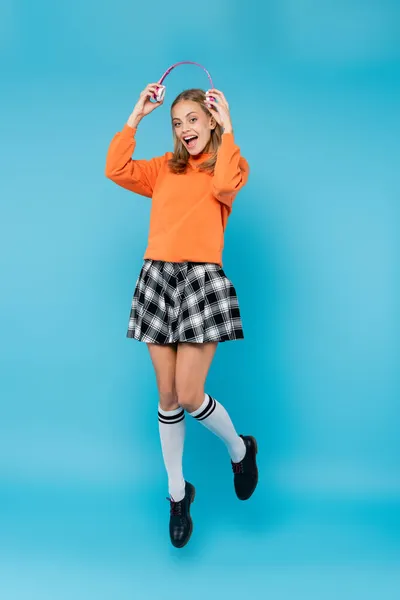 Cheerful student holding headphones while jumping on blue background — Stock Photo