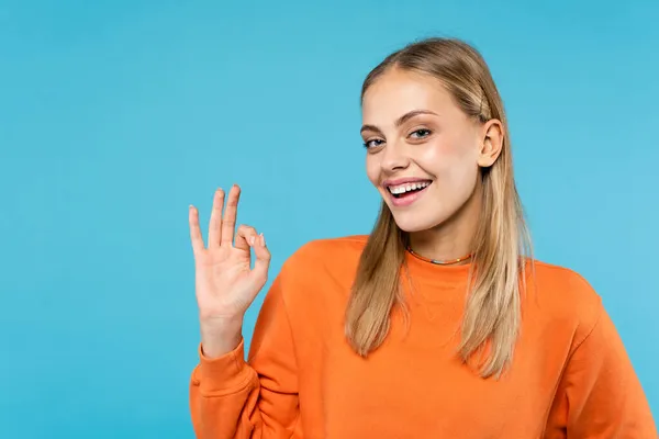 Cheerful woman in orange sweatshirt showing ok gesture isolated on blue — Stock Photo