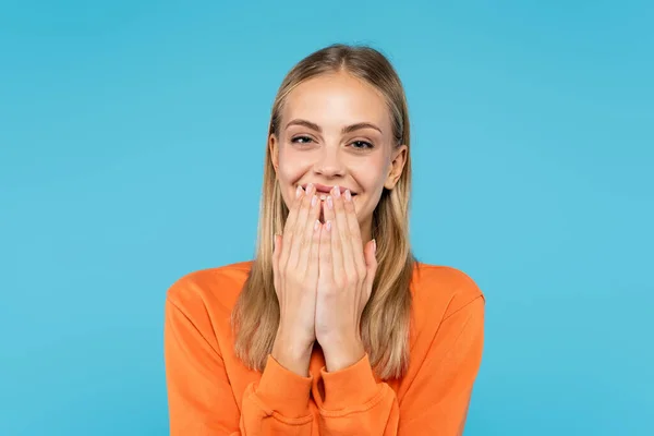 Glückliche blonde Frau bedeckt Mund mit Händen isoliert auf blau — Stockfoto