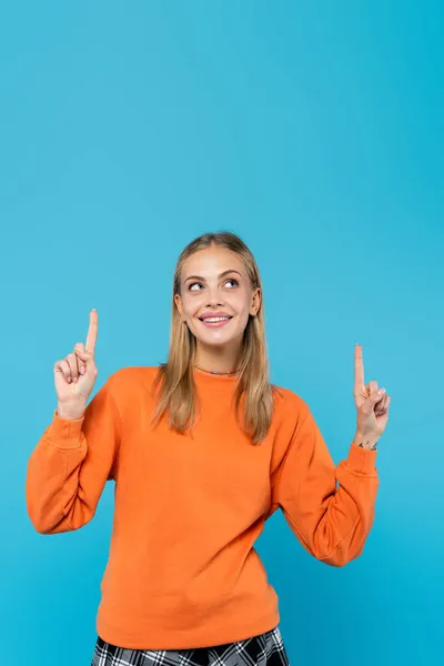 Smiling blonde woman in orange sweatshirt pointing with fingers isolated on blue — Stock Photo