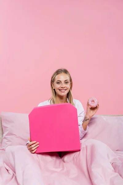 Femme blonde souriante en pyjama tenant beignet et boîte à pizza sur lit isolé sur rose — Photo de stock