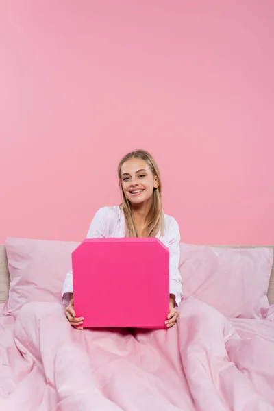 Happy blonde woman holding pizza box on bed isolated on pink — Stock Photo