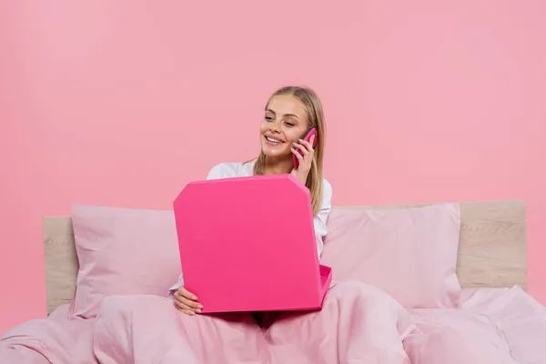 Smiling woman in pajamas talking on smartphone and holding pizza box on bed isolated on pink — Stock Photo