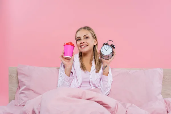 Femme blonde positive en pyjama tenant réveil et café pour aller isolé sur rose — Photo de stock