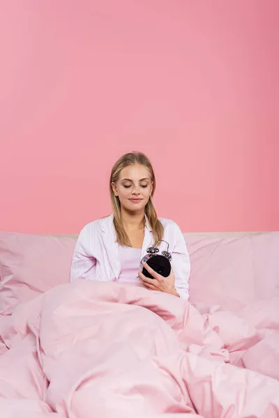 Young woman looking at alarm clock on bed isolated on pink — Stock Photo
