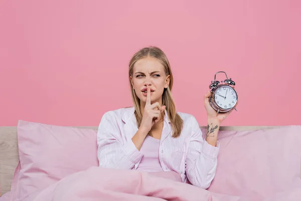 Mulher loira mostrando gesto secreto e segurando despertador na cama isolado em rosa — Fotografia de Stock