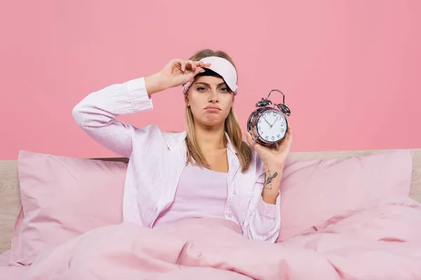 Mujer molesta sosteniendo la máscara del sueño y despertador en la cama aislado en rosa - foto de stock