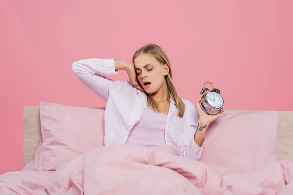 Blonde woman in pajamas holding alarm clock and yawning on bed isolated on pink — Stock Photo