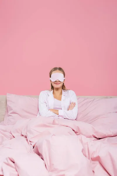 Femme souriante dans le masque de sommeil assis avec les bras croisés sur le lit isolé sur rose — Photo de stock