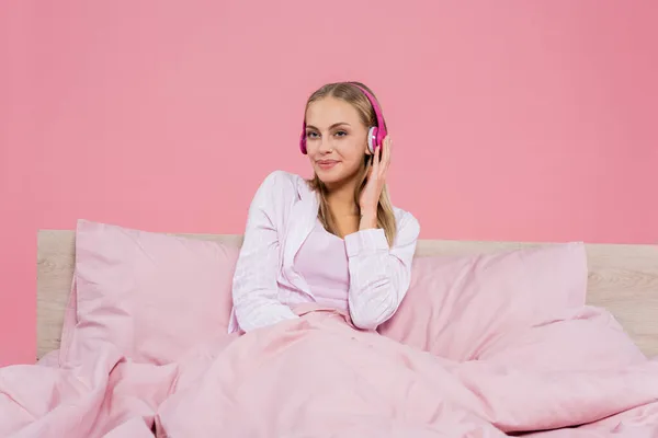 Jeune femme blonde en pyjama écoutant de la musique dans des écouteurs sur le lit isolé sur rose — Photo de stock