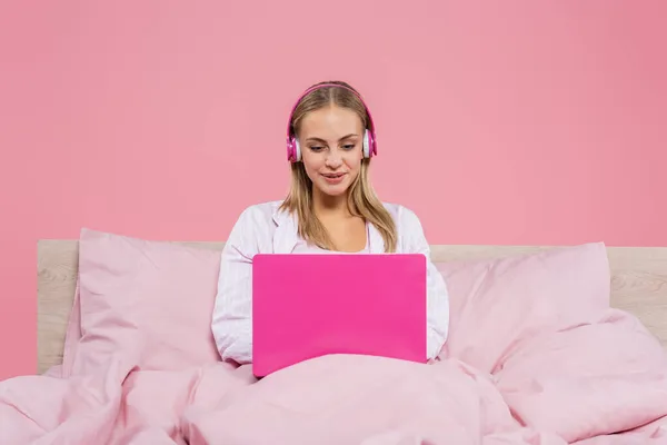 Freelance sorridente in cuffia utilizzando laptop sul letto isolato su rosa — Foto stock