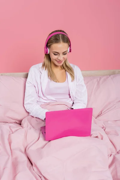 Donna positiva in cuffia e pigiama utilizzando computer portatile sul letto isolato su rosa — Foto stock