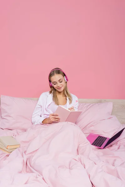 Freelancer in headphones writing on notebook near laptop and books on bed isolated on pink — Stock Photo