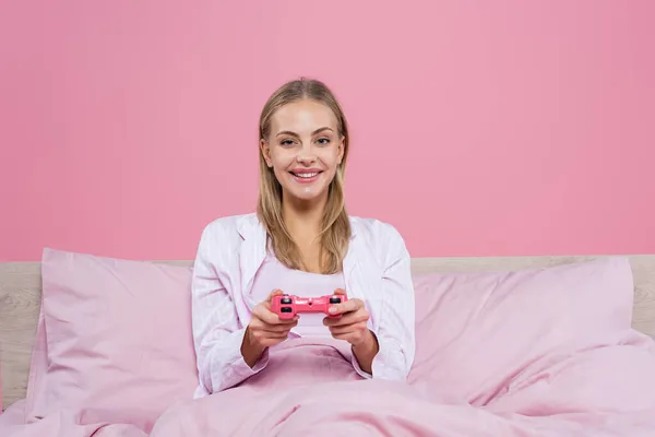 KYIV, UCRANIA - 27 de octubre de 2020: Mujer rubia positiva jugando videojuegos en la cama aislada en rosa - foto de stock