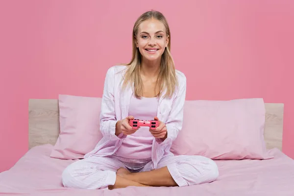 KYIV, UCRANIA - 27 de octubre de 2020: Mujer joven en pijama jugando videojuegos en la cama aislada en rosa - foto de stock