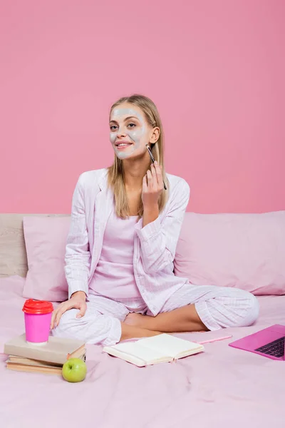 Femme souriante appliquant masque facial près d'un ordinateur portable et des livres sur le lit isolé sur rose — Photo de stock