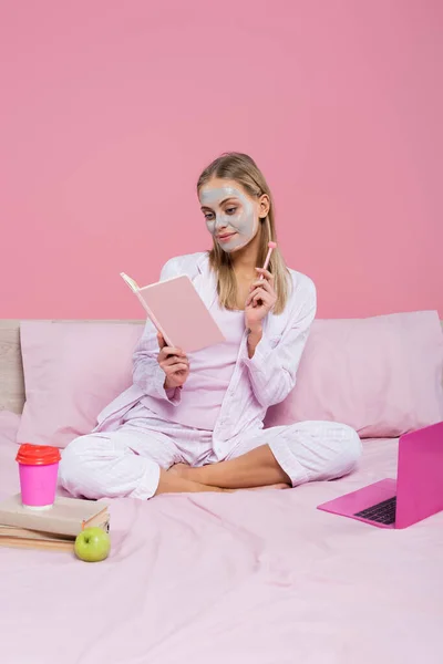 Blonde freelancer with face mask holding notebook near books, laptop and paper cup on bed isolated on pink — Stock Photo