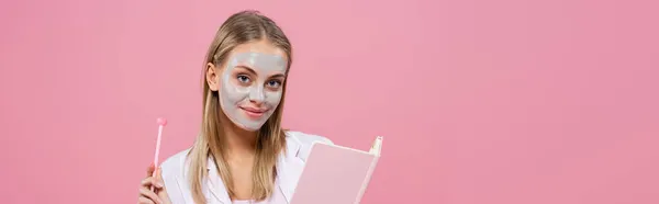 Blonde woman in pajamas and clay mask on face holding pen and notebook isolated on pink, banner — Stock Photo