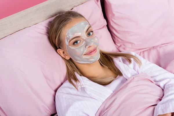 Vista de ángulo alto de la mujer rubia sonriente con máscara de arcilla en la cara mirando a la cámara en la cama aislada en rosa - foto de stock