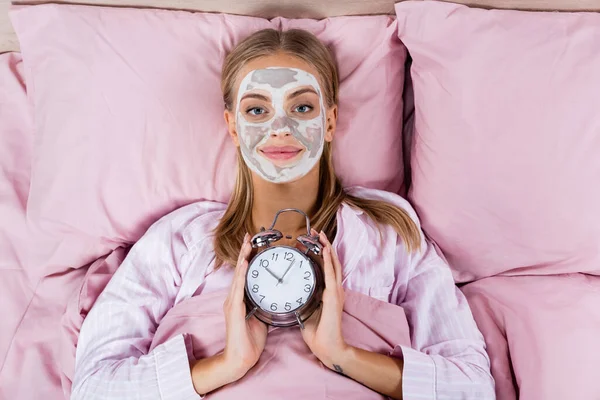 Draufsicht der lächelnden Frau mit Lehmmaske im Gesicht, die Wecker auf dem Bett hält, isoliert auf rosa — Stockfoto