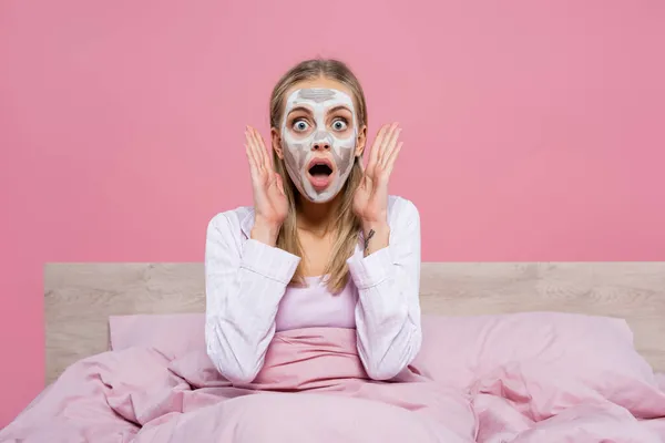 Donna scioccata con maschera di argilla sul viso guardando la fotocamera sul letto isolato su rosa — Stock Photo