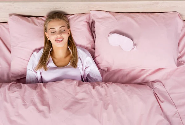 Vista superior de la mujer rubia que sobresale de la lengua cerca de la máscara del sueño en la cama - foto de stock