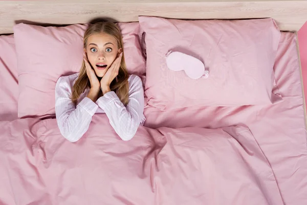Draufsicht der aufgeregten Frau im Pyjama mit Blick auf die Kamera auf dem Bett isoliert auf rosa — Stockfoto