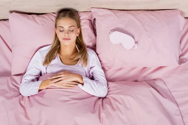 Vue du dessus de la femme blonde dormant près du masque sur le lit — Photo de stock